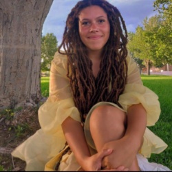 Image of Nate Combs, Secretary/Social Media Marketing Officer of Black Student Association. The image portrays a young person with long, thick, brown dreadlocks sitting on grass near a large tree trunk. The individual is wearing a light yellow, sheer, and flowy dress. The person has a relaxed, content expression and is sitting with their hands clasped around their knees, which are drawn up loosely in front of them. In the background, there is an expanse of green grass and several leafy trees under a clear blue sky, reflecting a sunny day.